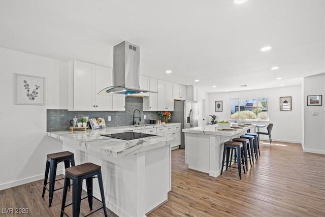 kitchen with black electric stovetop, stainless steel fridge with ice dispenser, island range hood, wood finished floors, and a kitchen bar