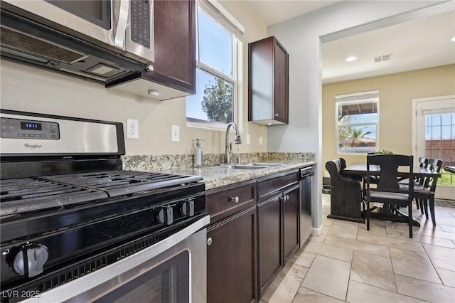 kitchen with dark brown cabinetry, light tile patterned floors, appliances with stainless steel finishes, light stone countertops, and a sink