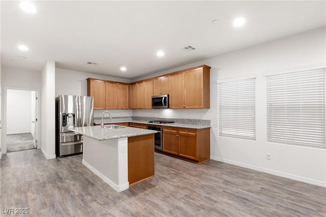 kitchen featuring stainless steel appliances, light stone countertops, a center island with sink, and light wood-style flooring