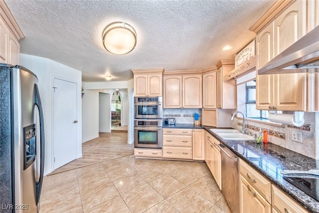 kitchen with backsplash, light brown cabinets, wall chimney range hood, appliances with stainless steel finishes, and a sink