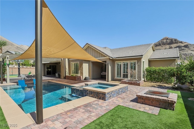 back of house with an outdoor fire pit, a patio, a pool with connected hot tub, a mountain view, and board and batten siding