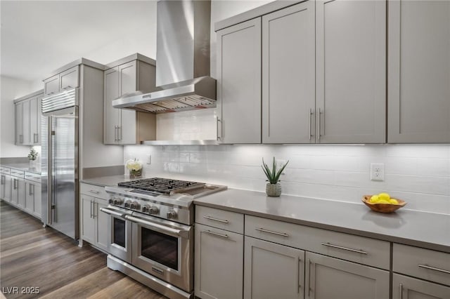 kitchen with high end appliances, tasteful backsplash, gray cabinets, wood finished floors, and wall chimney exhaust hood