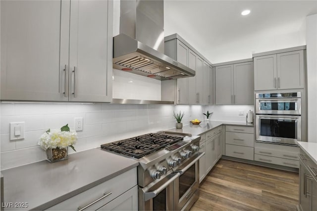 kitchen featuring dark wood-style floors, stainless steel appliances, gray cabinetry, wall chimney range hood, and backsplash