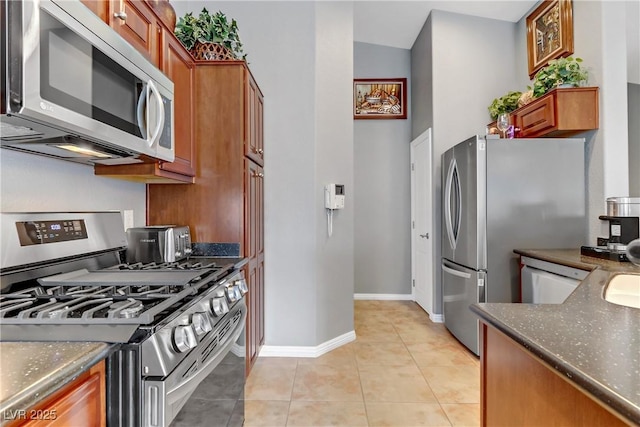 kitchen with light tile patterned floors, stainless steel appliances, baseboards, brown cabinets, and dark stone counters