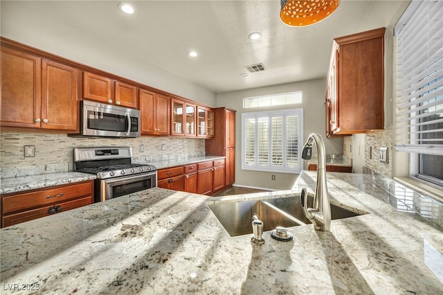 kitchen featuring light stone counters, stainless steel appliances, a sink, backsplash, and brown cabinets