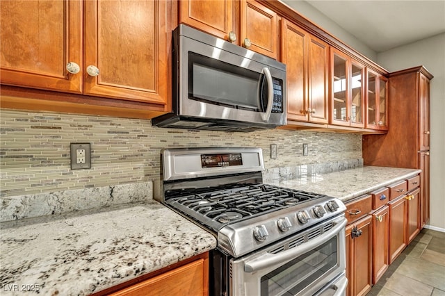 kitchen featuring decorative backsplash, glass insert cabinets, appliances with stainless steel finishes, brown cabinets, and light stone countertops