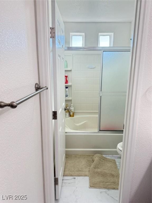 bathroom featuring a textured wall, marble finish floor, shower / bath combination with glass door, and toilet