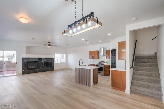 kitchen with a ceiling fan, stainless steel gas range oven, wall chimney range hood, brown cabinets, and light countertops