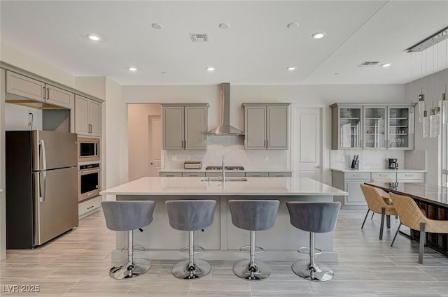 kitchen featuring a center island with sink, wall chimney exhaust hood, stainless steel appliances, light countertops, and gray cabinetry