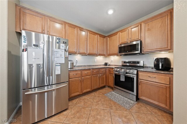 kitchen with appliances with stainless steel finishes, dark countertops, brown cabinetry, and light tile patterned floors