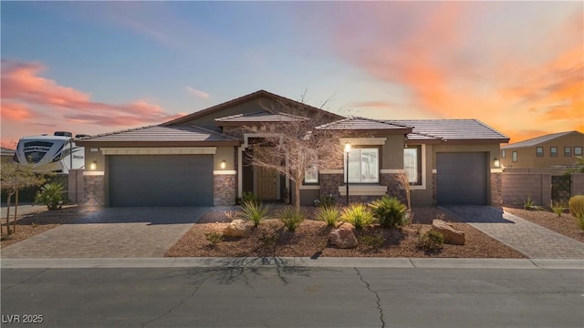 prairie-style house featuring an attached garage, fence, stone siding, decorative driveway, and stucco siding