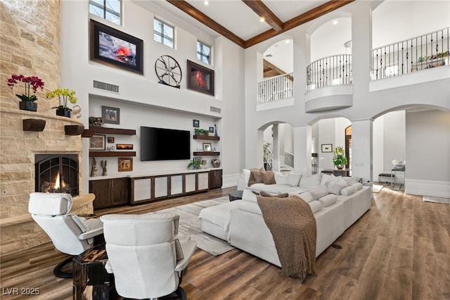 living room featuring visible vents, arched walkways, wood finished floors, a stone fireplace, and beam ceiling