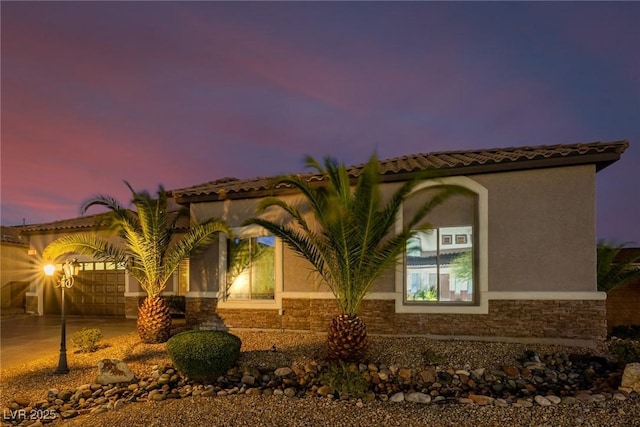 property exterior at dusk with a garage, stone siding, a tiled roof, driveway, and stucco siding