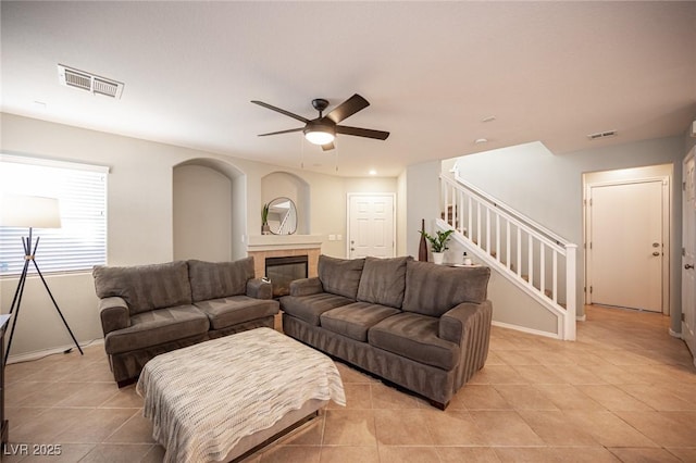 living area featuring a fireplace, visible vents, a ceiling fan, light tile patterned flooring, and stairs