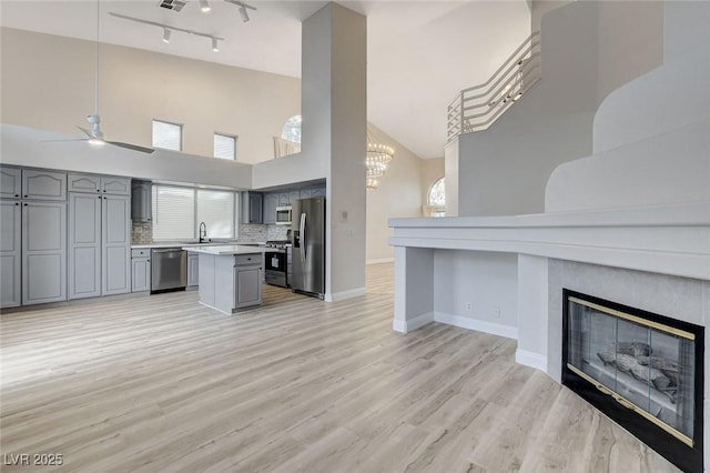 living room with high vaulted ceiling, ceiling fan with notable chandelier, a glass covered fireplace, light wood-style floors, and baseboards