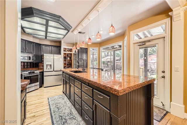 kitchen with a kitchen island with sink, light wood-style flooring, stainless steel appliances, a sink, and decorative light fixtures