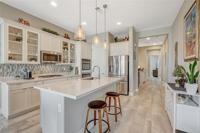 kitchen featuring decorative backsplash, glass insert cabinets, a kitchen island with sink, stainless steel appliances, and light countertops