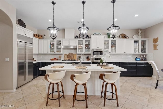 kitchen with built in appliances, a kitchen island with sink, a sink, white cabinets, and dark cabinetry