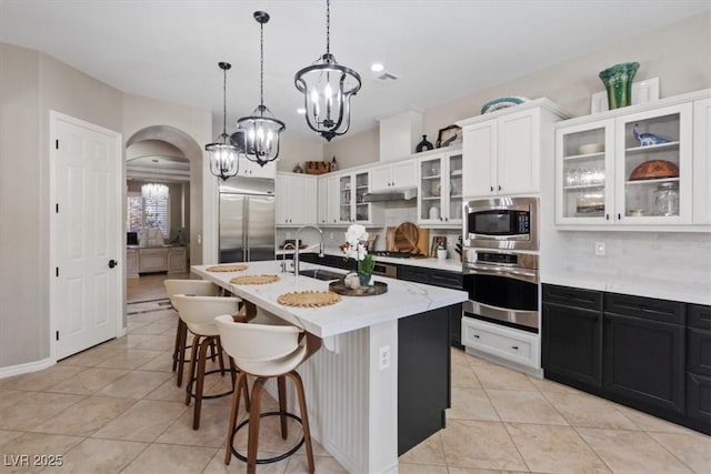 kitchen featuring arched walkways, a sink, built in appliances, dark cabinetry, and under cabinet range hood