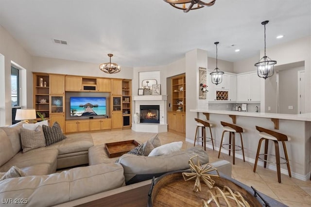 living area featuring a chandelier, light tile patterned flooring, visible vents, baseboards, and a glass covered fireplace