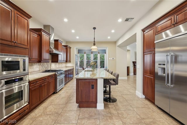 kitchen featuring light stone counters, a breakfast bar, built in appliances, wall chimney range hood, and backsplash