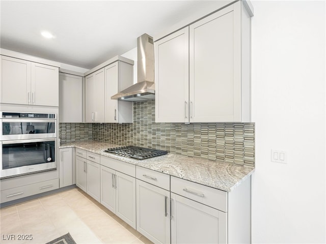 kitchen with appliances with stainless steel finishes, light stone counters, gray cabinetry, wall chimney range hood, and backsplash