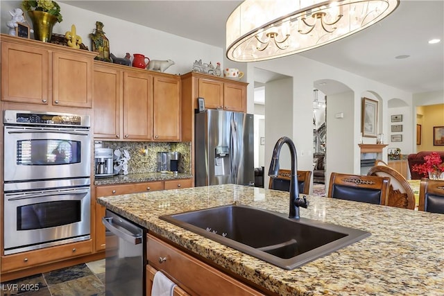 kitchen with a sink, appliances with stainless steel finishes, decorative backsplash, light stone countertops, and brown cabinetry