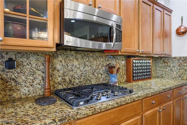 kitchen featuring light stone counters, stainless steel appliances, tasteful backsplash, brown cabinetry, and glass insert cabinets