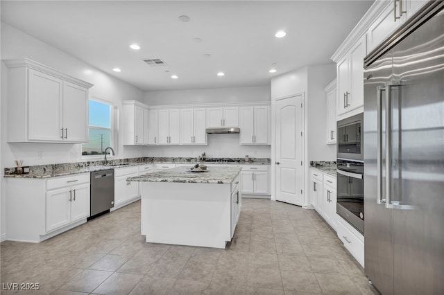 kitchen with white cabinets, a center island, built in appliances, light stone countertops, and under cabinet range hood