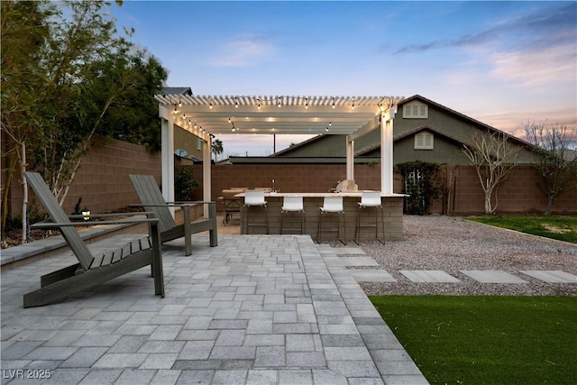 patio terrace at dusk with a fenced backyard, outdoor dry bar, an outdoor kitchen, and a pergola