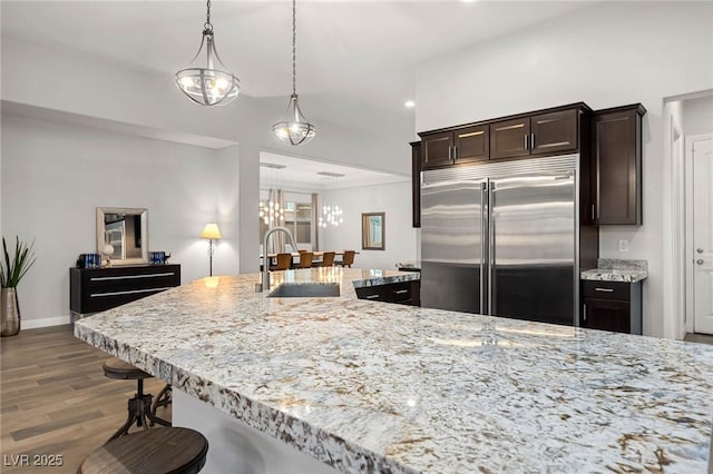 kitchen with dark brown cabinetry, built in refrigerator, light stone countertops, a kitchen bar, and a sink