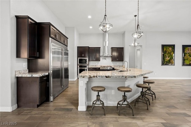 kitchen featuring built in appliances, dark brown cabinetry, a sink, a kitchen breakfast bar, and wall chimney exhaust hood