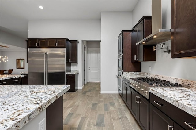 kitchen featuring dark brown cabinetry, baseboards, appliances with stainless steel finishes, wall chimney exhaust hood, and light wood finished floors