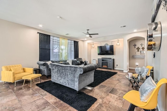 living area featuring visible vents, a ceiling fan, a warm lit fireplace, arched walkways, and baseboards