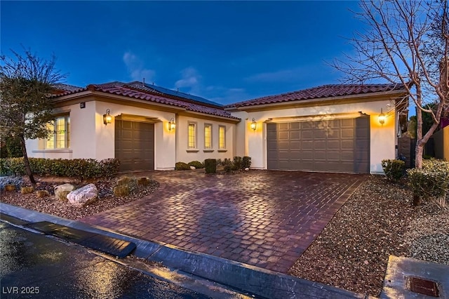 mediterranean / spanish-style house with decorative driveway, a tile roof, stucco siding, an attached garage, and roof mounted solar panels