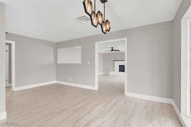 empty room featuring ceiling fan with notable chandelier, wood finished floors, visible vents, baseboards, and a brick fireplace