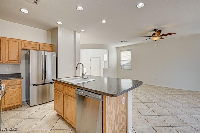 kitchen with light tile patterned floors, stainless steel appliances, dark countertops, a sink, and an island with sink