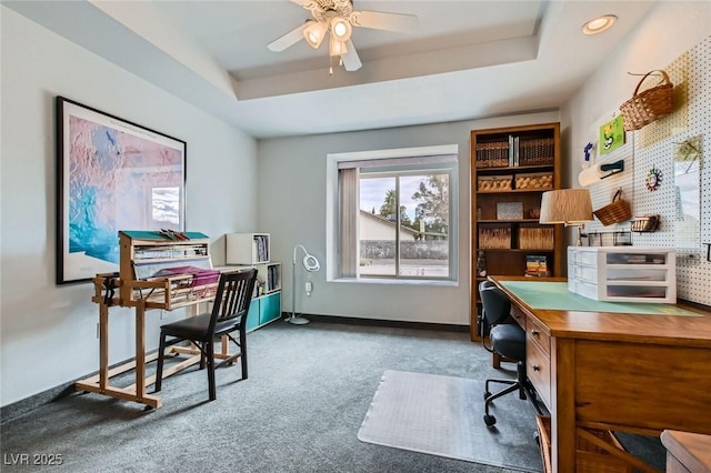 carpeted office space with a raised ceiling, a ceiling fan, and baseboards