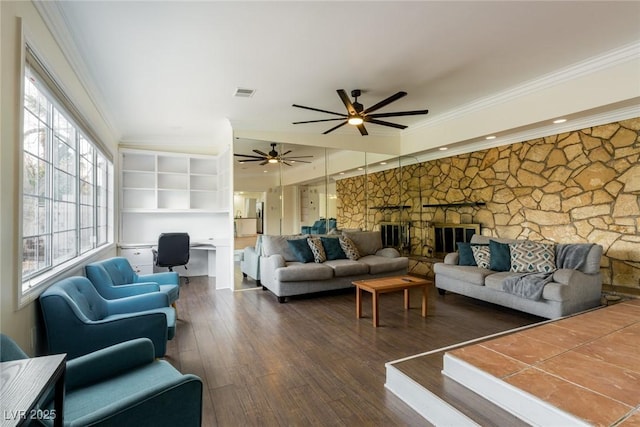 living room featuring built in desk, visible vents, ornamental molding, dark wood-type flooring, and a stone fireplace