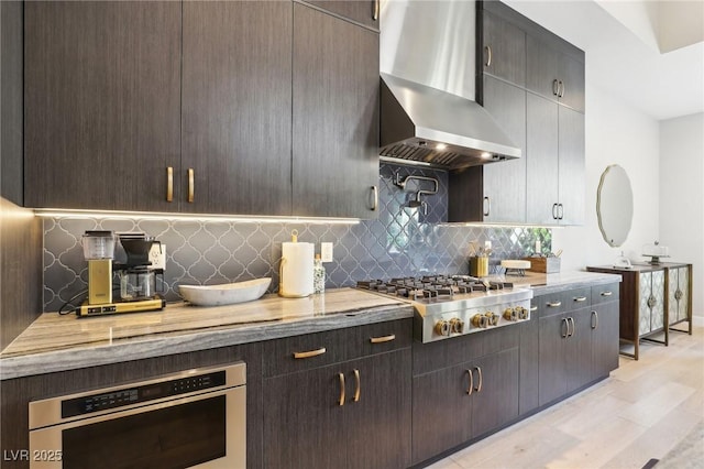 kitchen featuring dark brown cabinetry, light wood-style flooring, wall chimney range hood, appliances with stainless steel finishes, and decorative backsplash