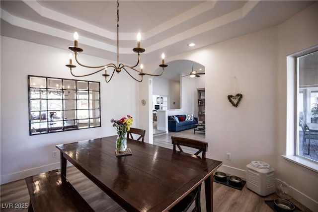 dining room with baseboards, arched walkways, wood finished floors, a tray ceiling, and ceiling fan with notable chandelier