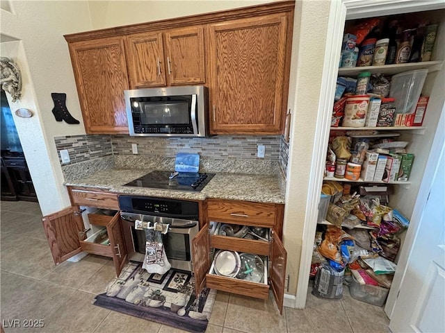 kitchen with light tile patterned floors, decorative backsplash, light stone counters, brown cabinets, and stainless steel appliances