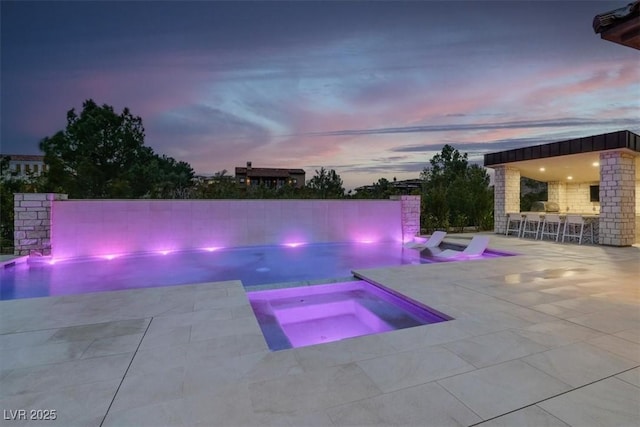 pool at dusk with a patio, a fenced backyard, outdoor wet bar, area for grilling, and a pool with connected hot tub