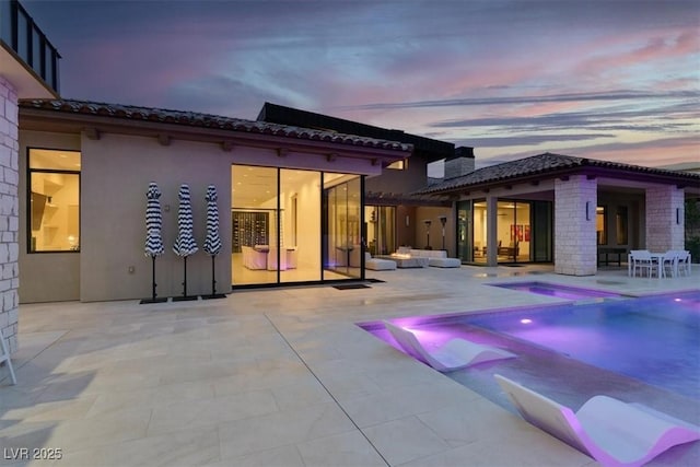 back of house at dusk featuring a patio area, a tile roof, a pool with connected hot tub, and stucco siding