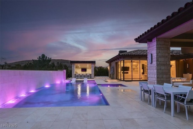 pool at dusk featuring a storage structure, an outdoor structure, outdoor dry bar, outdoor dining space, and a patio area