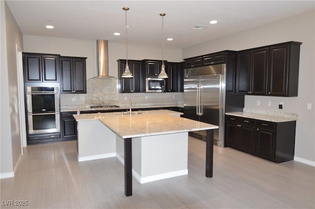 kitchen featuring appliances with stainless steel finishes, light stone countertops, a kitchen island with sink, wall chimney range hood, and a sink