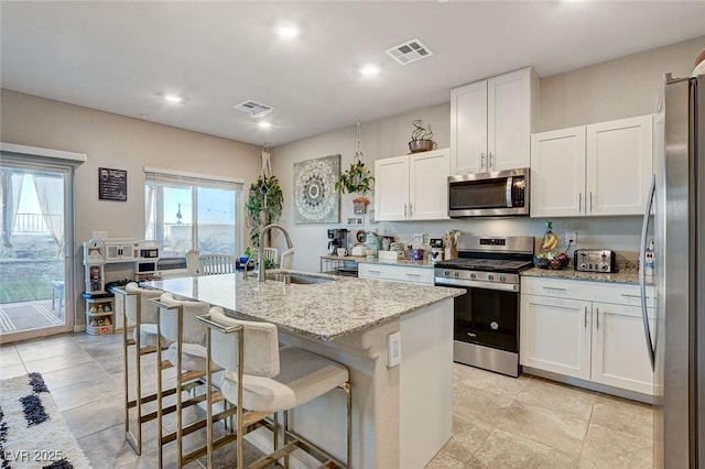 kitchen with visible vents, a kitchen island with sink, a sink, stainless steel appliances, and light stone countertops