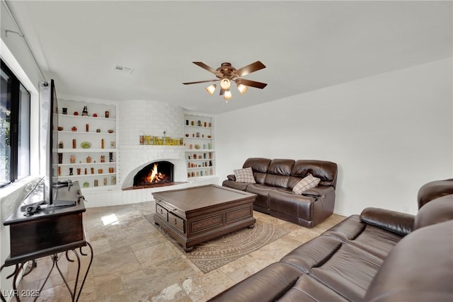 living room with ceiling fan, a fireplace, visible vents, built in features, and stone finish floor