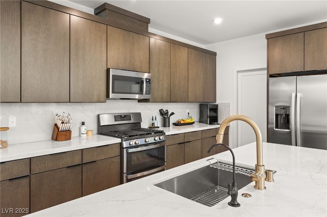 kitchen with light stone counters, dark brown cabinetry, stainless steel appliances, a sink, and backsplash