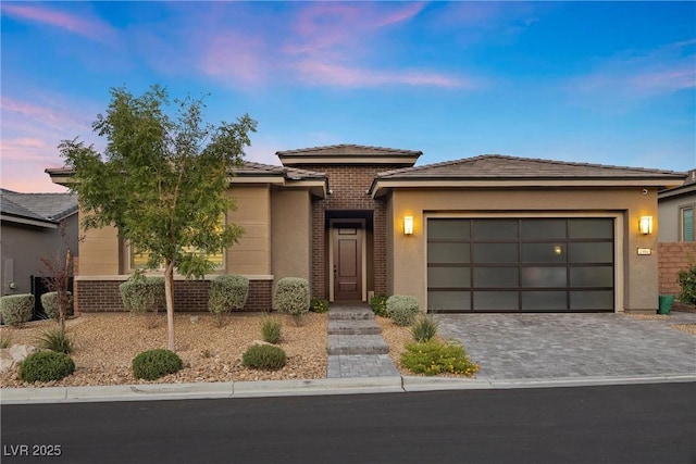 prairie-style house with a garage, brick siding, decorative driveway, and stucco siding